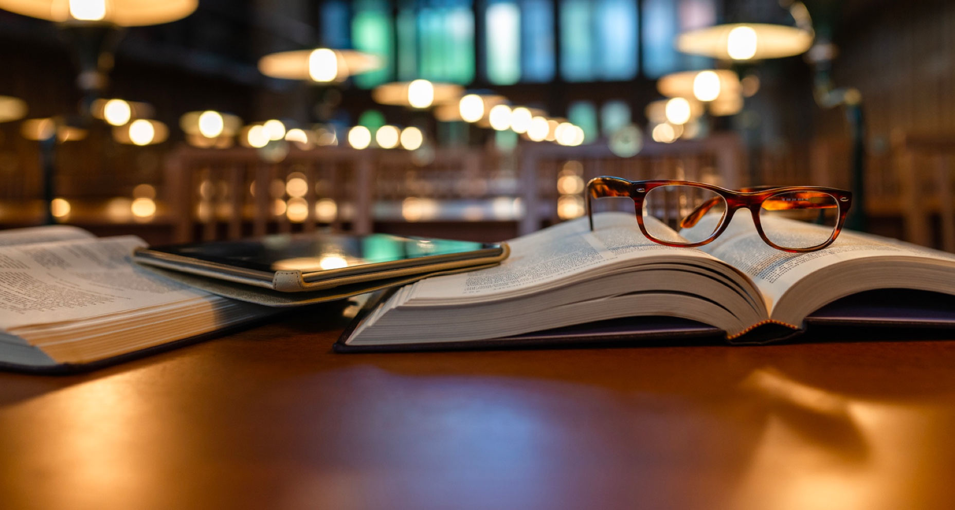 一副眼镜放在一本书上，电子平板电脑放在图书馆的桌子上。
